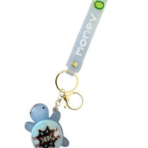 May include: A light blue turtle keychain with a gold-colored clasp and a light blue strap. The strap has the word "money" printed on it. The turtle has a decorative design on its shell.
