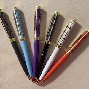 May include: Six pens with gold accents and patterned designs. The pens are in various colours, including black, blue, purple, white, and orange.