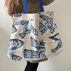May include: A beige tote bag with a blue bird pattern and blue handles. The bag features a repeating design of stylized birds in shades of blue against a neutral background. The handles are a solid, vibrant blue.