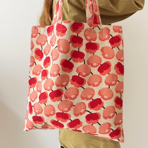May include: A beige tote bag featuring a repeating pattern of red and pink floral shapes. The bag has matching handles. The floral design is a mix of solid and detailed shapes. The bag is being held.