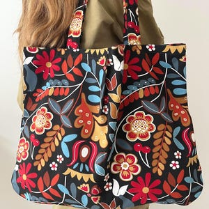 May include: A black tote bag with a colourful floral design. The pattern includes red, orange, yellow, and blue flowers and leaves on a black backdrop. The bag has matching handles and is carried over the shoulder.