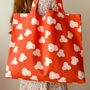 May include: A large, red tote bag with white floral patterns. The bag has matching red handles and is made of a sturdy fabric. The floral design is a repeating pattern of white flowers with yellow centers.