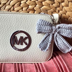 May include: A white leather crossbody bag with a silver MK logo and a gray crocheted bow charm.