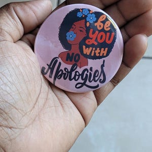 Pode incluir: Bot&atilde;o rosa com uma ilustra&ccedil;&atilde;o preta e marrom de uma mulher com cabelo afro e flores no cabelo. O texto no bot&atilde;o diz "Be you with no apologies".