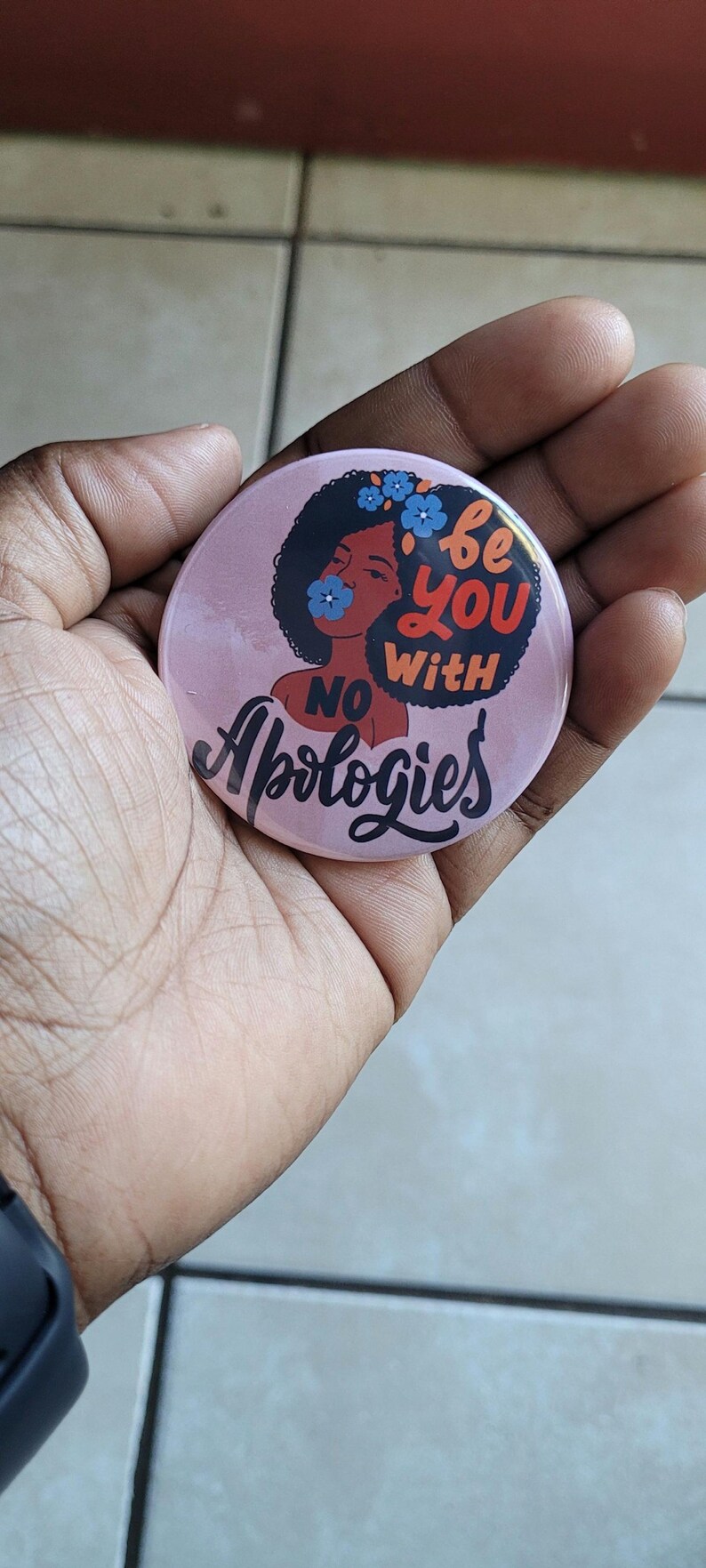 Pode incluir: Bot&atilde;o rosa com uma ilustra&ccedil;&atilde;o preta e marrom de uma mulher com cabelo afro e flores no cabelo. O texto no bot&atilde;o diz "Be you with no apologies".