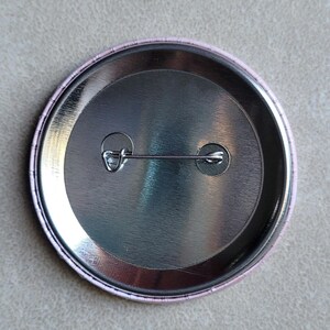 Pode incluir: A parte de tr&aacute;s de um bot&atilde;o redondo com um suporte de metal prateado e uma borda de pl&aacute;stico rosa. O bot&atilde;o tem um pino de seguran&ccedil;a preso &agrave; parte de tr&aacute;s.