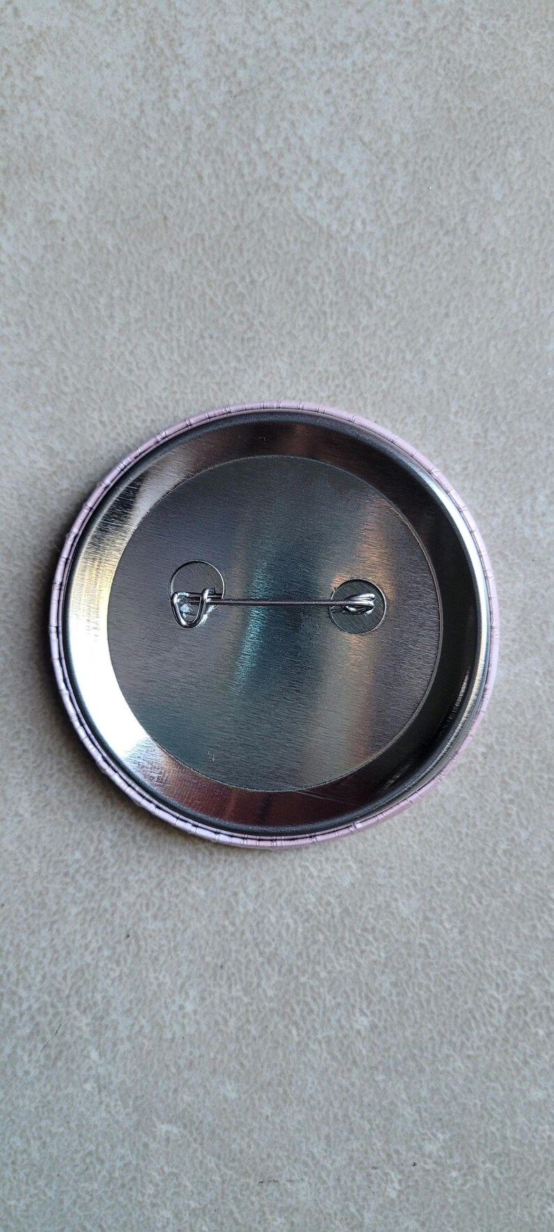 Pode incluir: A parte de tr&aacute;s de um bot&atilde;o redondo com um suporte de metal prateado e uma borda de pl&aacute;stico rosa. O bot&atilde;o tem um pino de seguran&ccedil;a preso &agrave; parte de tr&aacute;s.