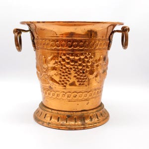 May include: A polished copper ice bucket featuring embossed grape clusters and vine leaf motifs on the body. The vessel includes two side handles with circular rings and a wide, flared base with a decorative border pattern around the rim.