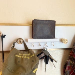 May include: A white wall-mounted organizer with a gold-toned frame, featuring four decorative knobs, a small shelf, and five metal hooks. A brown wallet rests on the shelf, while a green baseball cap, keys, and a plaid fabric item hang from the knobs.