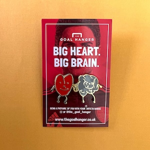 Könnte beinhalten: Zwei Emaille-Pins mit einem roten Herzen und einem grauen Gehirn, beide mit Beinen und Armen. Die Pins befinden sich auf einer roten und weißen Karte mit dem Text "THE GOAL HANGER BIG HEART. BIG BRAIN." und "SEND A PICTURE OF YOU WITH YOUR ARTETA BADGE @the_goal_hanger adicwww.thegoalhanger.co.uk"