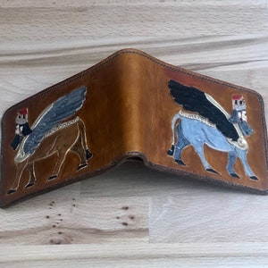 Puede incluir: Una cartera de cuero marrón con una imagen detallada de un toro alado a cada lado. El toro está representado en tonos de gris, negro y dorado.