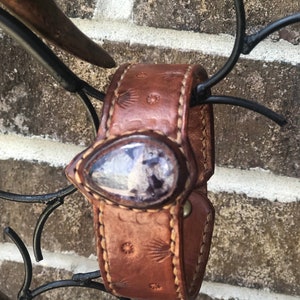Könnte beinhalten: Braunes Lederarmband mit einem dekorativen Edelstein. Das Armband hat detaillierte Nähte und geprägte Muster. Der Edelstein ist in Tropfenform gefasst. Das Armband ist an einem dekorativen schwarzen Metallstück befestigt.