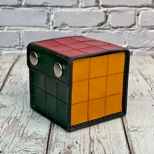 Può includere: Una piccola scatola quadrata in pelle con un motivo a scacchi rosso, verde e giallo. La scatola ha due bottoni a pressione argentati sul davanti.
