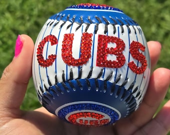 Op de afbeelding: Een witte baseball met blauwe en rode stiksels. Het woord "CUBS" is gespeld in rode strass steentjes. De baseball heeft een blauw en rood logo aan de zijkant.