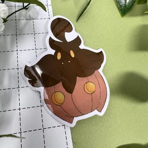 May include: A sticker featuring a cartoon character with a brown head and a pumpkin-shaped body. The character has yellow eyes and two yellow circles on the pumpkin body. The sticker is on a white background with a grid pattern.
