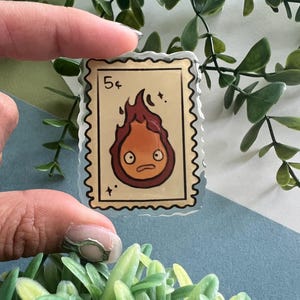 May include: A clear, rectangular sticker resembling a postage stamp, featuring a cartoon fire demon with a surprised expression. The stamp reads "5&cent;" in the upper left corner. The fire demon is red and orange with a brown outline.