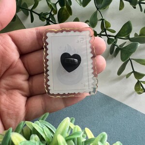 May include: A clear, rectangular pin with a gold border and a black heart-shaped pin back. The pin has a white background with a faint design and the number "2" in the upper right corner. The pin is held in a hand.
