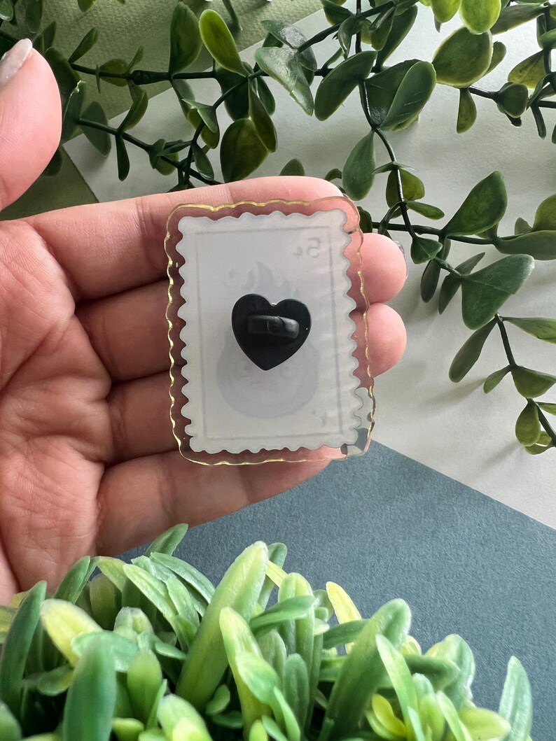 May include: A clear, rectangular pin with a gold border and a black heart-shaped pin back. The pin has a white background with a faint design and the number "2" in the upper right corner. The pin is held in a hand.