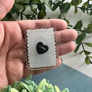 May include: A clear acrylic pin shaped like a postage stamp with a black heart design. The pin has a scalloped edge and a black pin back. The background is a mix of green foliage and a blue surface.