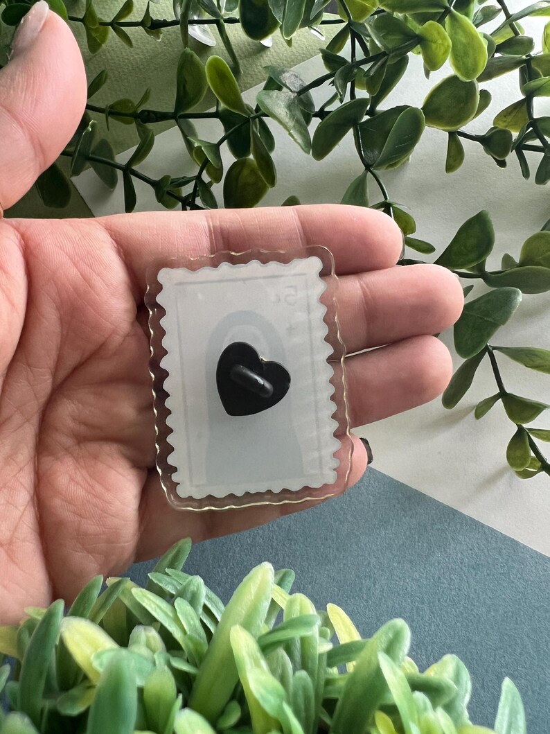 May include: A clear acrylic pin shaped like a postage stamp with a black heart design. The pin has a scalloped edge and a black pin back. The background is a mix of green foliage and a blue surface.
