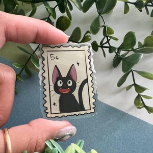 May include: A clear sticker featuring a black cat illustration in a postage stamp design. The stamp reads "5&cent;" and includes a black cat with large eyes and a curved tail. The sticker is held against a backdrop of green foliage.