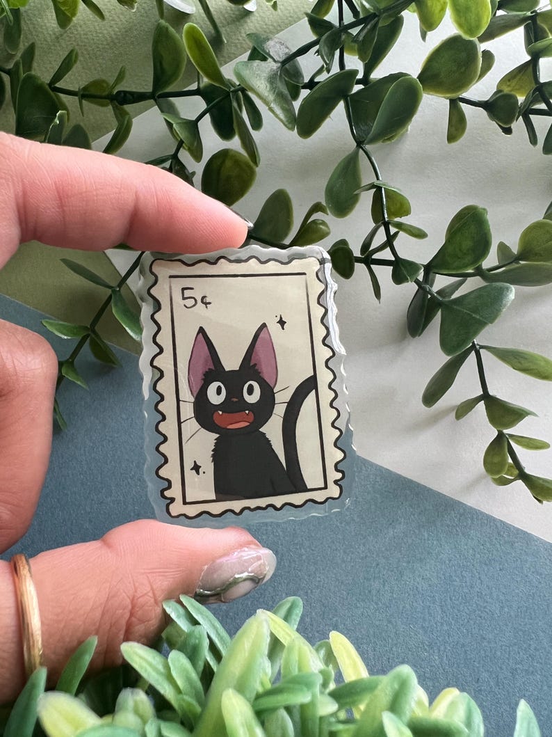 May include: A clear sticker featuring a black cat illustration in a postage stamp design. The stamp reads "5&cent;" and includes a black cat with large eyes and a curved tail. The sticker is held against a backdrop of green foliage.