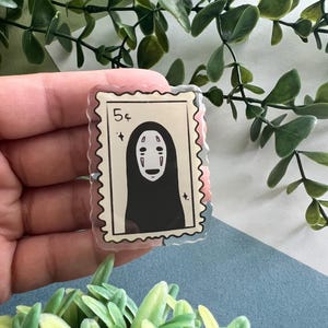 May include: A clear acrylic stamp-shaped pin featuring a black and white illustration of a character with a blank face. The pin has a 5&cent; marking and a jagged edge, resembling a postage stamp. The background includes green foliage and a blue surface.