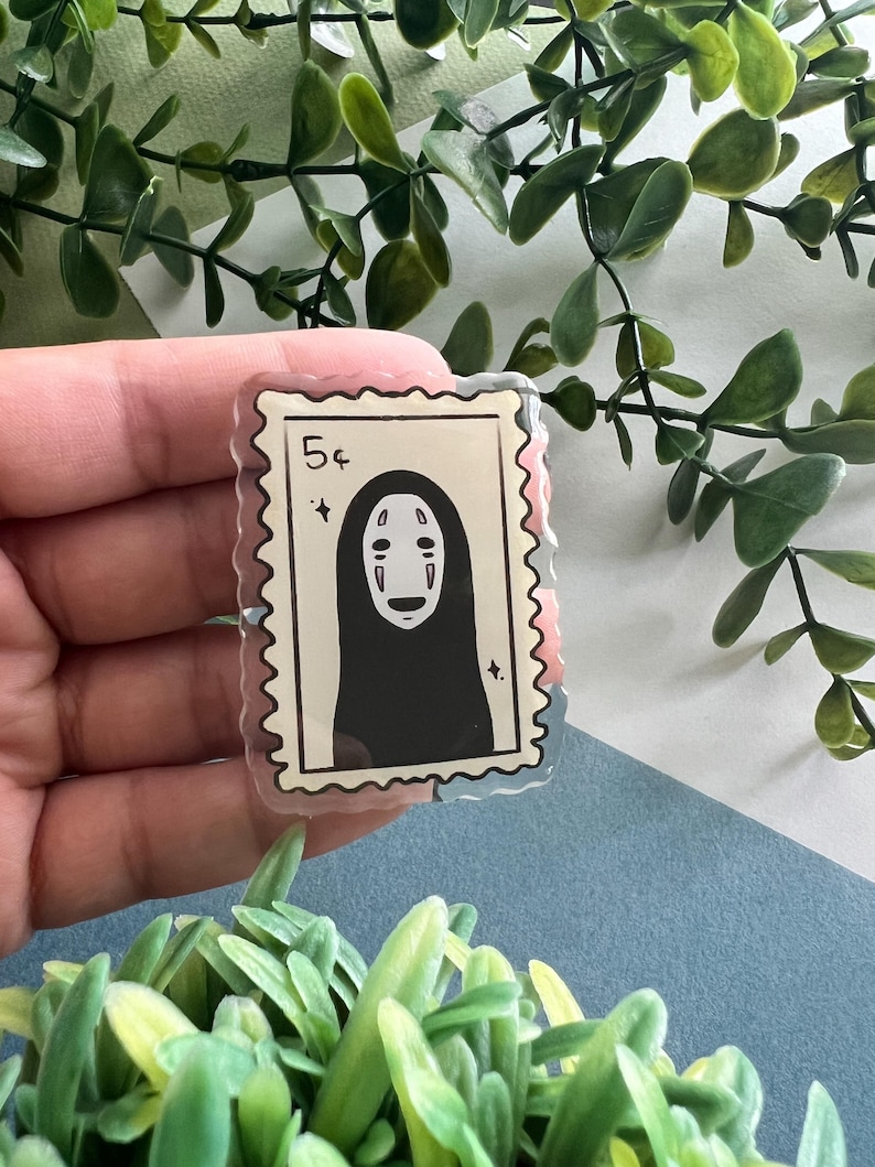 May include: A clear acrylic stamp-shaped pin featuring a black and white illustration of a character with a blank face. The pin has a 5&cent; marking and a jagged edge, resembling a postage stamp. The background includes green foliage and a blue surface.