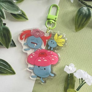 Peut inclure: Un porte-clés en acrylique transparent avec des personnages de dessins animés en forme de champignon bleus, roses et jaunes. Le porte-clés a un fermoir vert et est entouré de feuillage vert et de fleurs blanches.