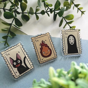May include: Three clear acrylic pins in the shape of postage stamps. Each pin features a different cartoon character: a black cat, a fire demon, and a faceless spirit. The pins are outlined with a light blue border and the price "5&cent;".