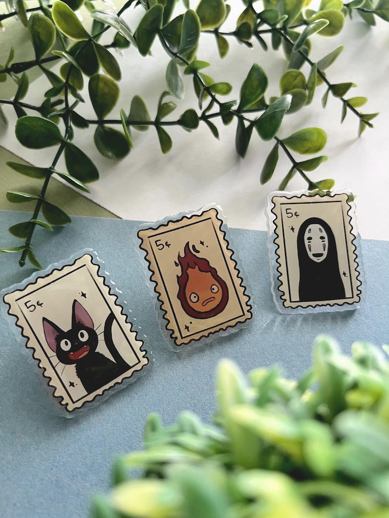 May include: Three clear acrylic pins in the shape of postage stamps. Each pin features a different cartoon character: a black cat, a fire demon, and a faceless spirit. The pins are outlined with a light blue border and the price "5&cent;".