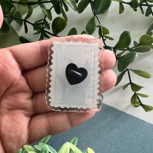 May include: A clear acrylic pin with a postage stamp design. The pin features a black heart-shaped backing and a light gray cat silhouette. The edges of the pin are scalloped, and the text "$2" is visible.