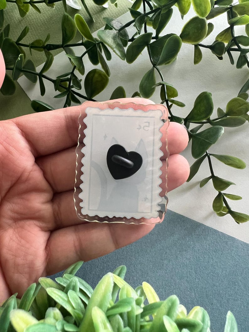 May include: A clear acrylic pin with a postage stamp design. The pin features a black heart-shaped backing and a light gray cat silhouette. The edges of the pin are scalloped, and the text "$2" is visible.