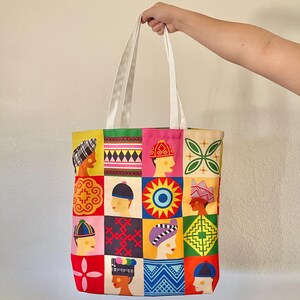 May include: A colorful tote bag with a patchwork design featuring various patterns and illustrations of people wearing different head coverings. The bag has white handles and a vibrant mix of colors including red, yellow, green, and pink.