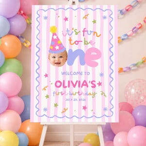 Può includere: Un cartello di compleanno rettangolare su un cavalletto bianco con sfondo a righe rosa e bianche e bordo blu smerlato. Il testo recita: it's fun to be one, WELCOME TO OLIVIA'S first birthday e JULY 20, 2026, con la foto di un bambino.