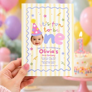 Op de afbeelding: Een rechthoekige uitnodigingskaart voor een eerste verjaardag met gele verticale strepen en een golvende paarse rand. Het bevat een foto van een baby met een feesthoedje, de tekst 'it's fun to be one', 'Olivia's First Birthday' en evenementdetails.