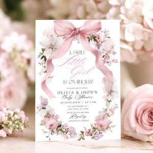 以下が含まれることがあります： ベビーシャワーの招待状。花輪のデザインで、ピンクのリボンと「A Sweet Little Girl Is On Her Way！」の文字が特徴です。オリビアとジョンズのベビーシャワーの詳細が含まれています。