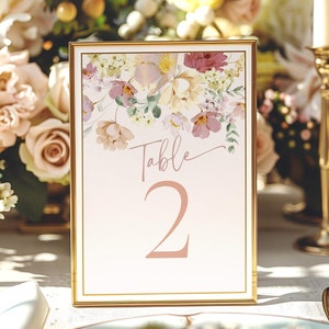 May include: A framed table number with a gold border. The table number is white with a floral design at the top and the word "Table" and the number "2" in a soft pink color. The background includes flowers and a candle.