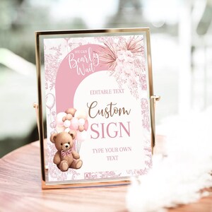 Pode incluir: Um sinal imprimível rosa e marrom com um urso de pelúcia segurando balões. O sinal diz "We can Bearly Wait" e "Custom Sign".