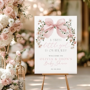 May include: A baby shower sign on a wooden easel, featuring a pink bow and floral design. The sign reads "A sweet little girl is on her way!" and welcomes guests to Olivia & Jhon's Baby Shower, dated 02.18.2025. The background includes floral arrangements.