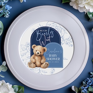May include: A white plate with a blue and white round paper plate on top. The paper plate has a blue border with the text "We can Bearly Wait" and a teddy bear illustration. The text "Caroline's Baby Shower 05.12.2024" is also on the paper plate.