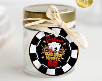 May include: A black and white casino chip with gold accents. The chip has a red and gold design with the text "Casino Night 50" and a playing card design. The chip is tied with a beige ribbon to a clear glass jar with a gold lid.