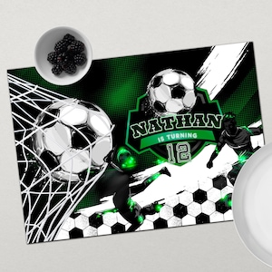 Op de afbeelding: Een groene en zwarte tafelrunner met een voetbalthema met een witte voetbal in een net, een voetballer die een bal schopt en de tekst "Nathan wordt 12".