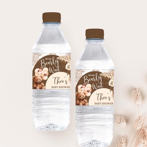 以下が含まれることがあります： 茶色のラベルが付いた透明なプラスチック製の水筒が2本。ラベルには、風船を持った漫画のテディベアと「We can Bearly Wait Theo's Baby Shower」という文字が書かれています。