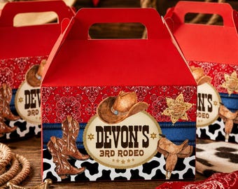 Puede incluir: Cajas de regalo rojas con temática de vaquero. Cada caja presenta una parte superior roja y un diseño con un sombrero de vaquero, botas y el texto "Devon's 3rd Rodeo". Las cajas están decoradas con un estampado de bandana y de piel de vaca.