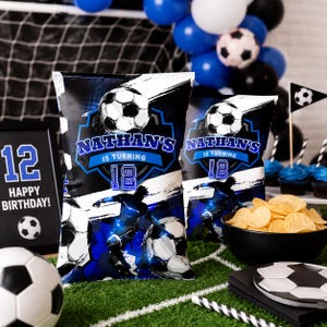 May include: A football-themed birthday party scene with black and blue decorations. Two crisp packets with "Nathan's is turning 12" text are displayed. A black bowl of crisps, cupcakes, and a football complete the scene.