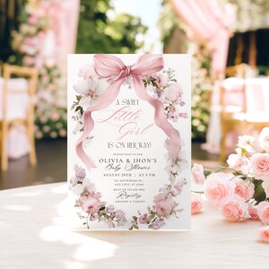 May include: A baby shower invitation with a floral wreath design. The invitation features the text "A Sweet Little Girl Is On Her Way!" and details for Olivia & Jhon's baby shower, including the date, time, and location. The invitation is decorated with pink flowers and a pink ribbon.
