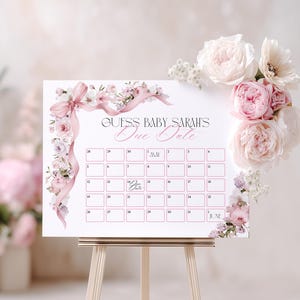 May include: A white baby shower calendar with the text "Guess Baby Sarah's Due Date" in elegant script. The calendar is decorated with pink floral accents and a pink ribbon. A bouquet of pink and white flowers adorns the top right corner.