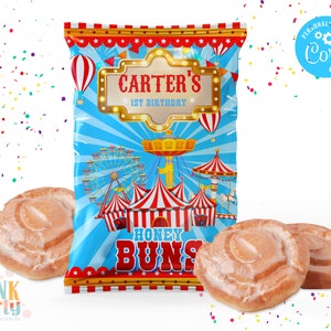 K&ouml;nnte beinhalten: Eine bunte Party-Geschenkt&uuml;te mit Jahrmarkt-Thema, mit Riesenrad, Zirkuszelten und Hei&szlig;luftballons. Auf der T&uuml;te steht "Carter's 1st Birthday" und "Honey Buns". Drei glasierte Honigbr&ouml;tchen liegen vor der T&uuml;te.