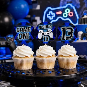 May include: Three cupcakes with white frosting, each topped with a circular blue and black gaming-themed topper. The toppers feature the text "GAME ON!", the name "JOSHUA" with a game controller graphic and the number "13", and the number "13".